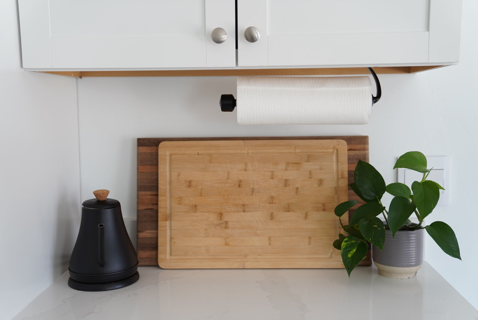 Wooden cutting board on a kitchen counter with a plant and black container.