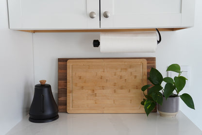 Wooden cutting board on a kitchen counter with a plant and black container.